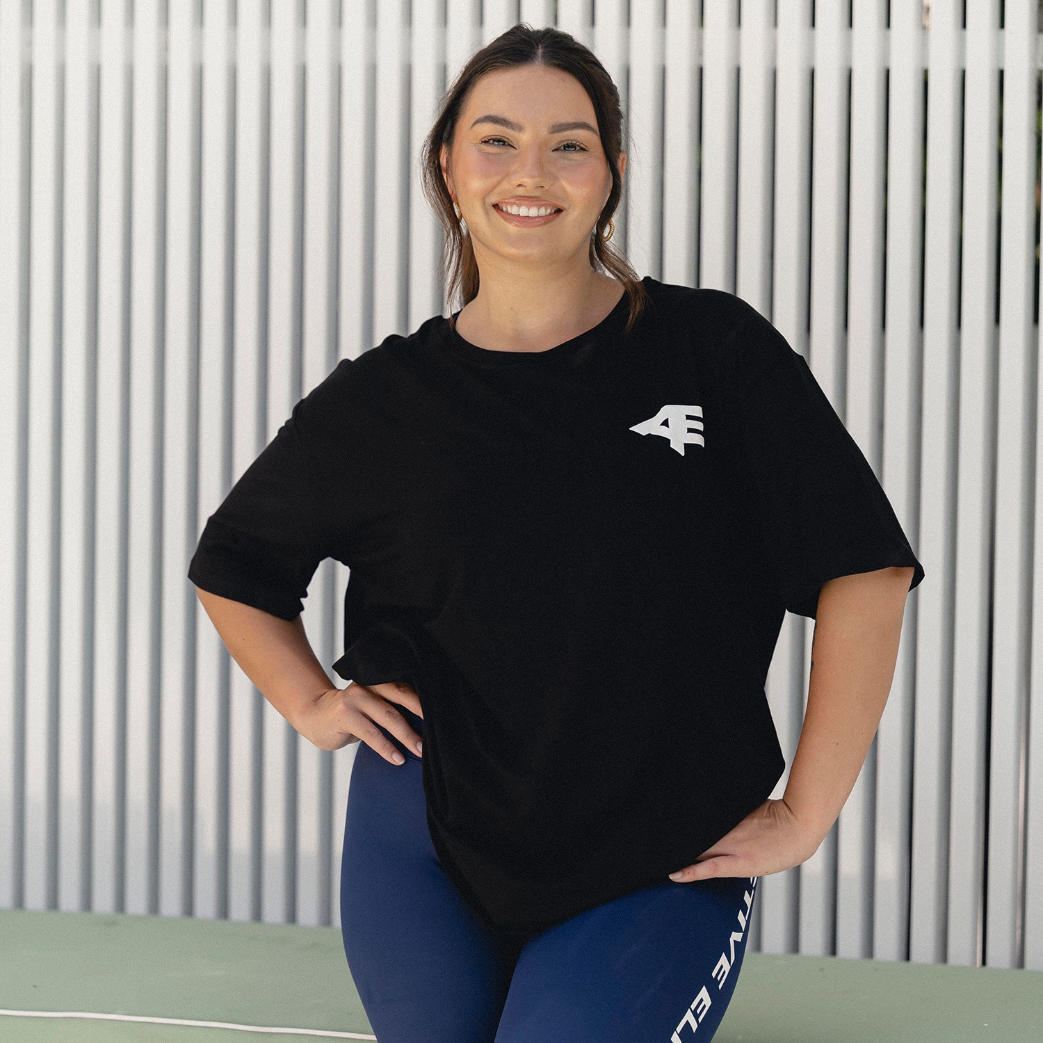 Smiling woman wearing a black Active Elite oversized tee and blue leggings, standing confidently in front of a modern white and green backdrop.