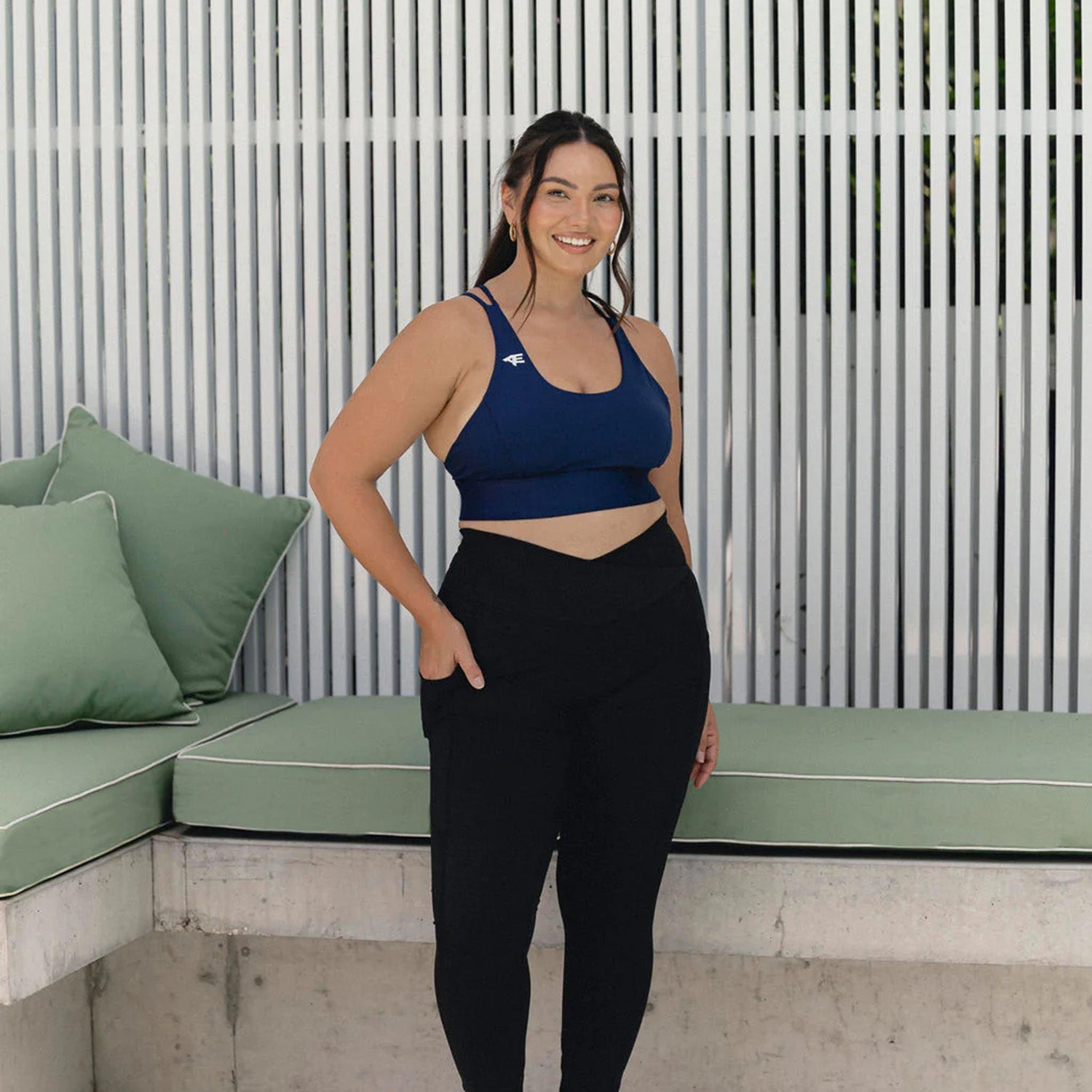 Smiling woman wearing Active Elite's navy sports bra and black Motivator Leggings, featuring a supportive V-shaped waistband and deep pockets, posing confidently by a modern outdoor lounge area.
