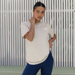 A woman wearing an oversized sandy coloured Active Elite t-shirt, standing in front of a light backdrop with her hand casually adjusting the shirt. She pairs the shirt with navy leggings, exuding a relaxed and confident vibe.
