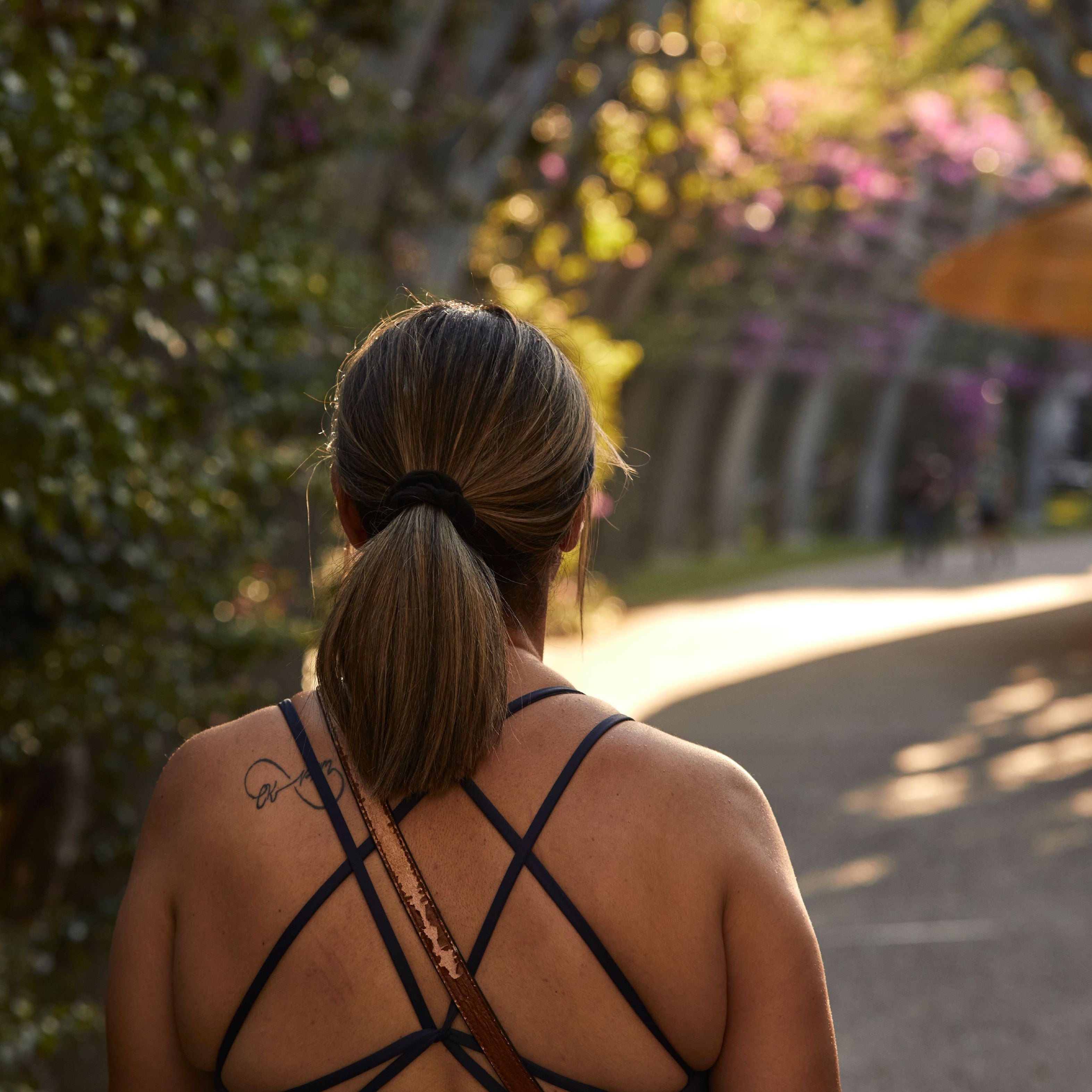 A woman captured from behind, wearing a black strappy sports bra, walking in a peaceful, nature-filled setting.