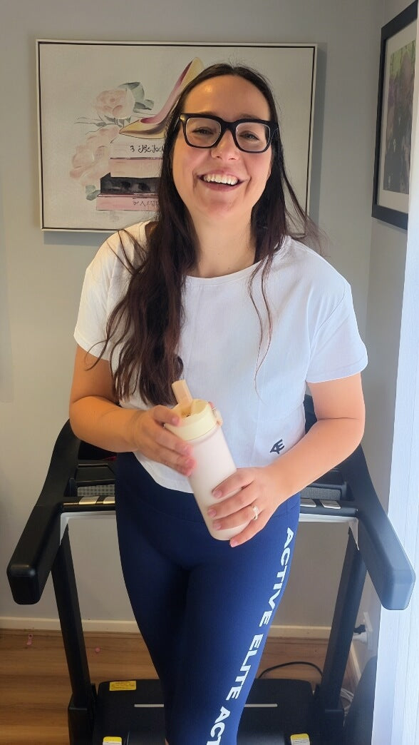 A woman on a treadmill in her activewear appearing happy and smiling and holding a water bottle.