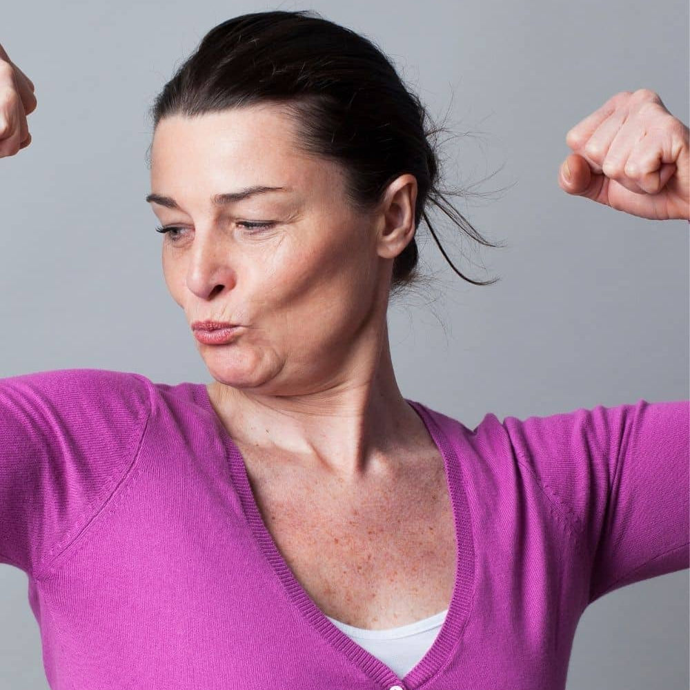 Woman aged 40+ flexing. She is wearing a plain purple cardigan and stood in front of a grey backdrop