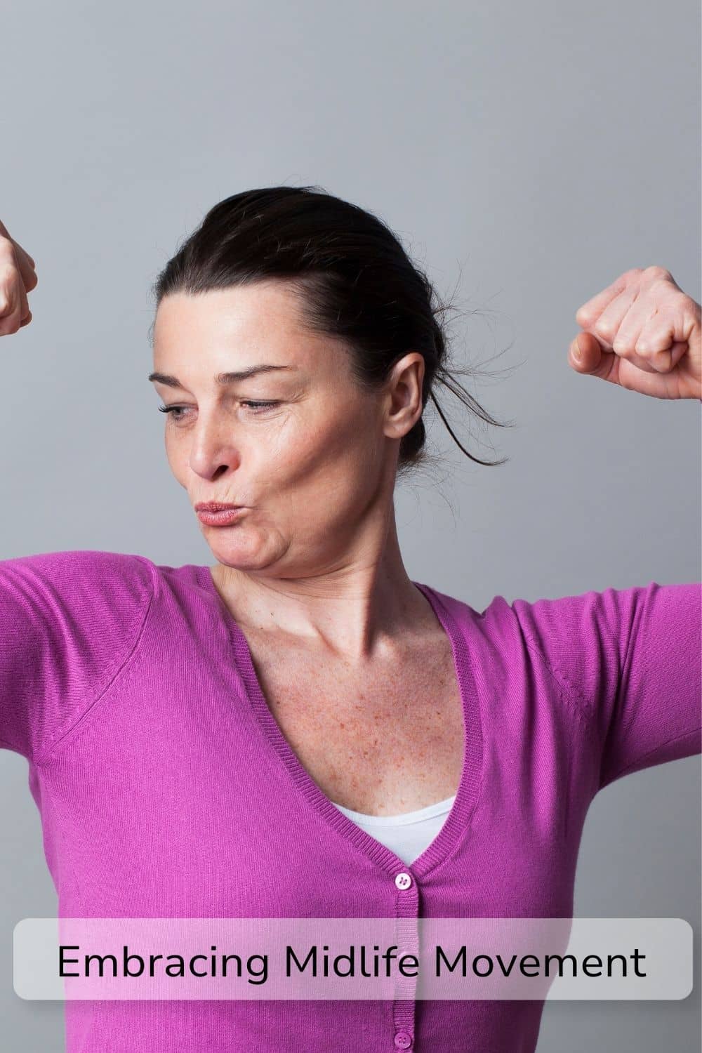 Woman aged 40+ flexing. She is wearing a plain purple cardigan and stood in front of a grey backdrop