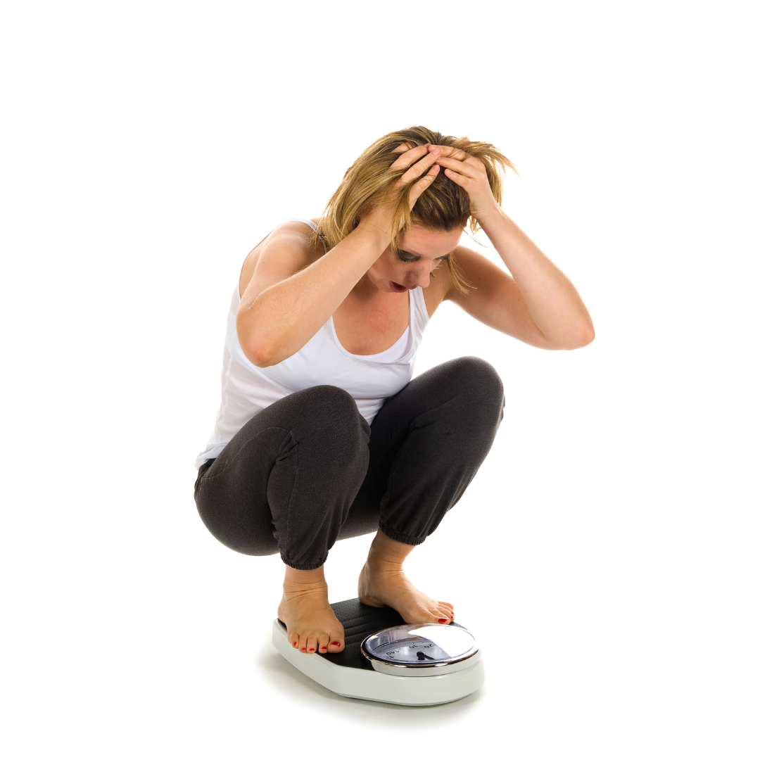 A woman crouching on a bathroom scale with her head in her hands, expressing frustration or concern about her weight.