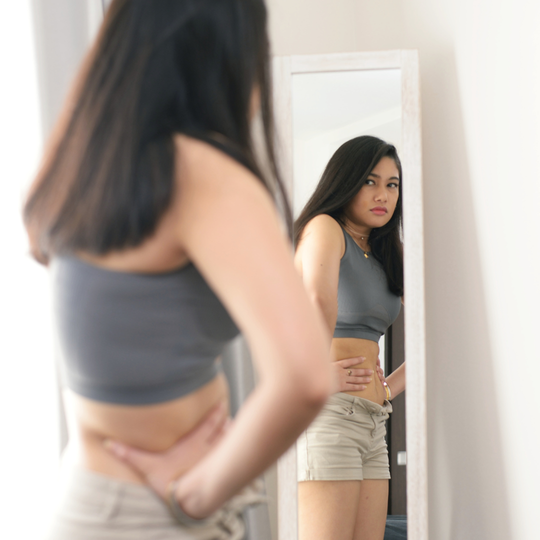 A young woman in a grey crop top and shorts standing in front of a full-length mirror, examining her reflection with a thoughtful, slightly concerned expression.