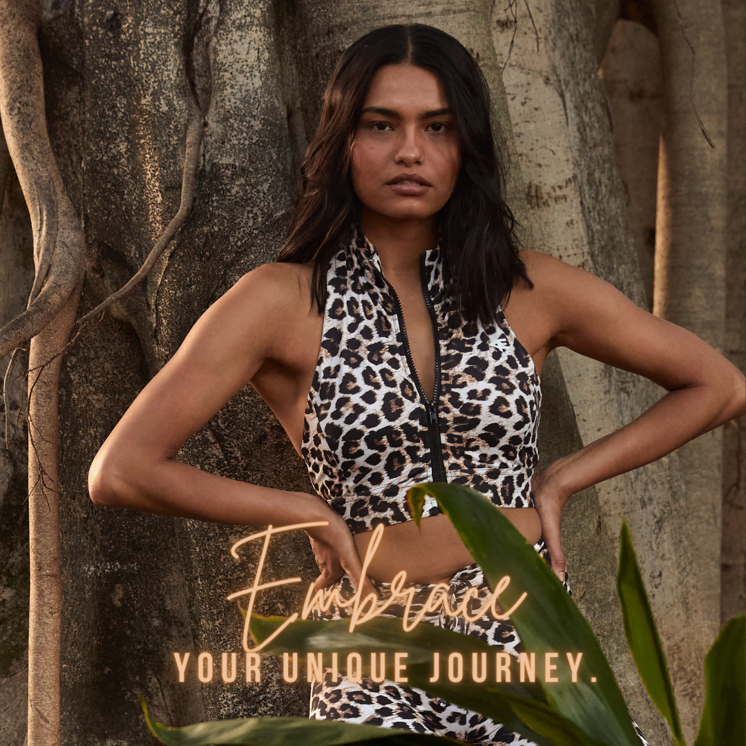 A woman standing against a natural background, arms crossed, wearing a leopard print sports bra with the text 'Embrace Your Unique Journey.'